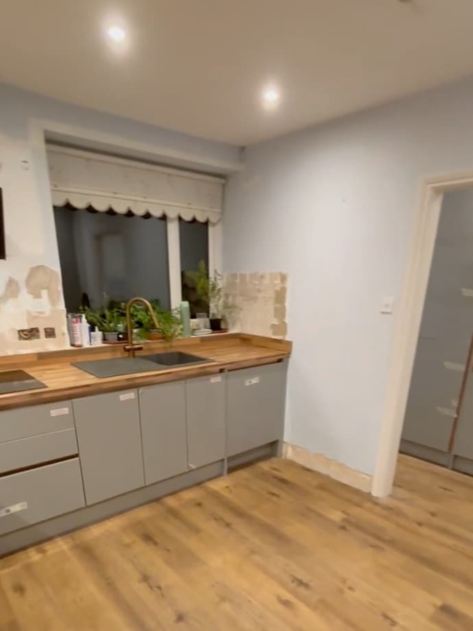 Finished work: a solid timber worktop with an inset sink and a brass tap, fitted grey units below it, and a new oak-effect floor running across the room.